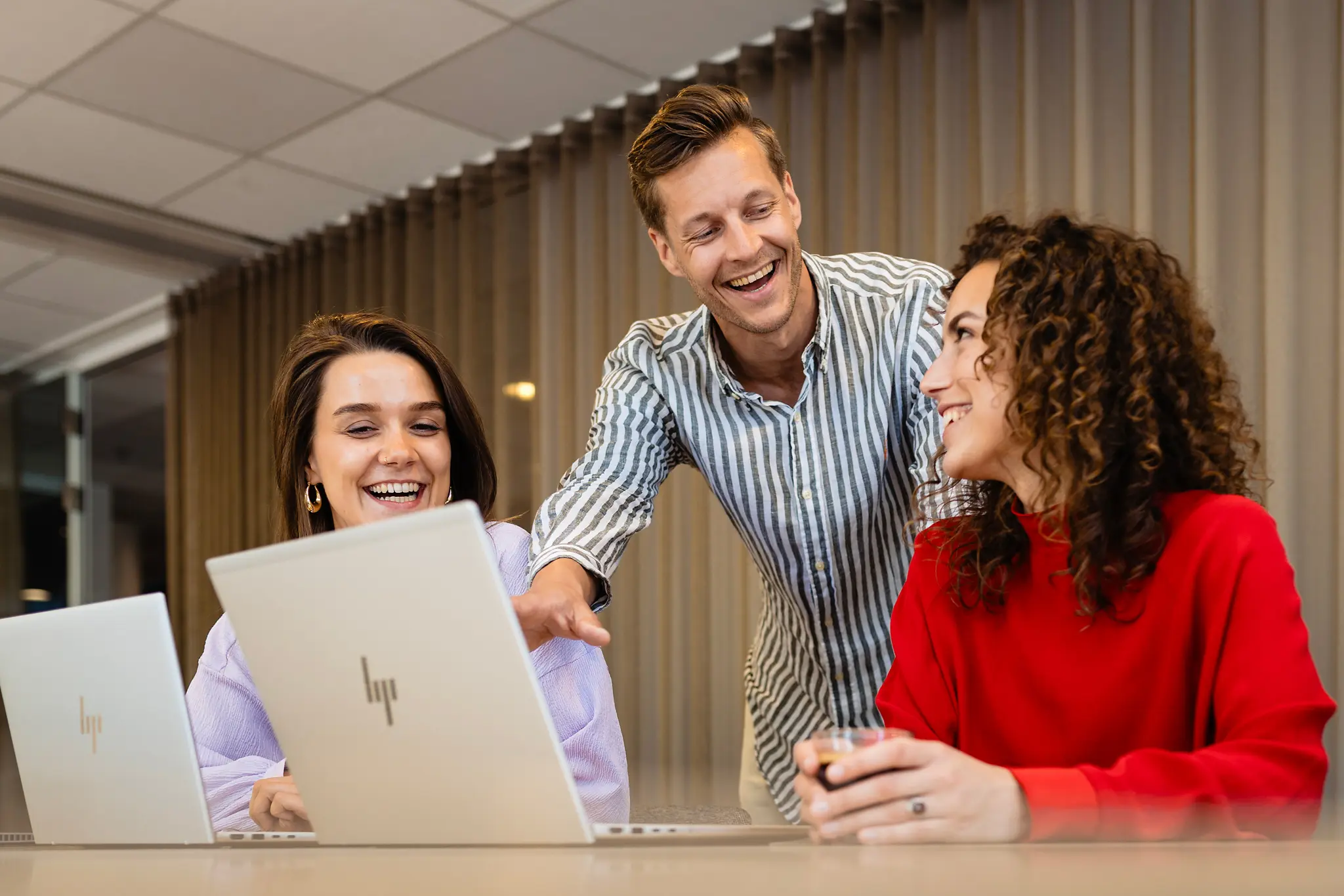 Drie mensen lachend, samenwerkend aan laptops in een kantooromgeving.