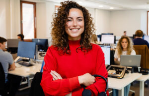 Vrouw in rode trui lacht in een kantoor met computers en collega's op de achtergrond.