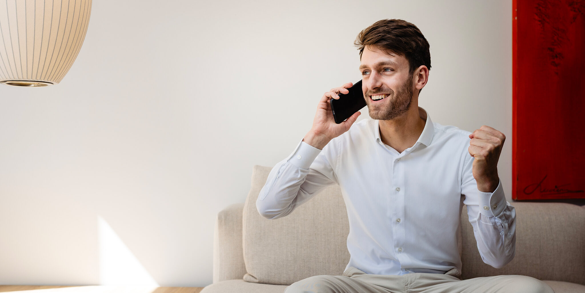 Man in een witte blouse zit op de bank en telefoneert met een glimlach en opgetrokken vuist.
